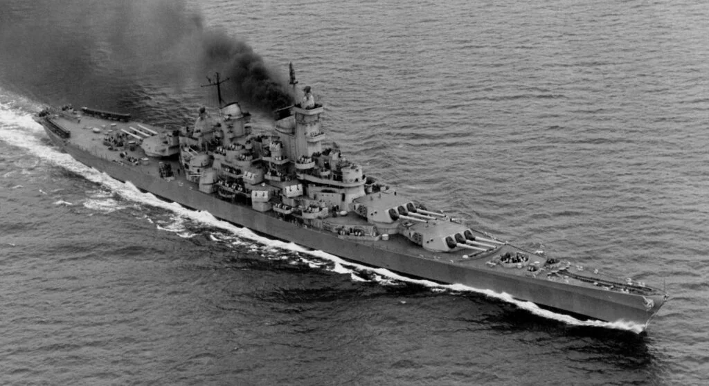 A large battleship sails through the ocean, trailing black smoke. Steeped in history, the ship boasts multiple large gun turrets and a complex superstructure, creating white waves as it cuts through the water. A large battleship sails through the ocean, trailing black smoke. Steeped in history, the ship boasts multiple large gun turrets and a complex superstructure, creating white waves as it cuts through the water.