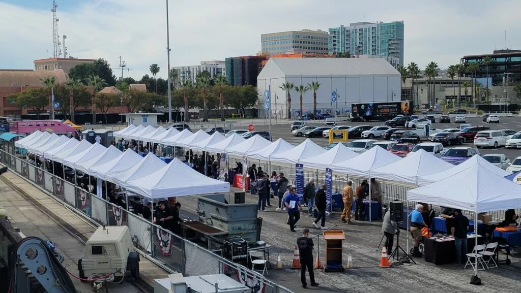 Rows of white tents line a parking lot, transforming the space into a vibrant event venue where people gather for an outdoor occasion. Cars are parked nearby, and tall buildings stand in the background under a partly cloudy sky.
