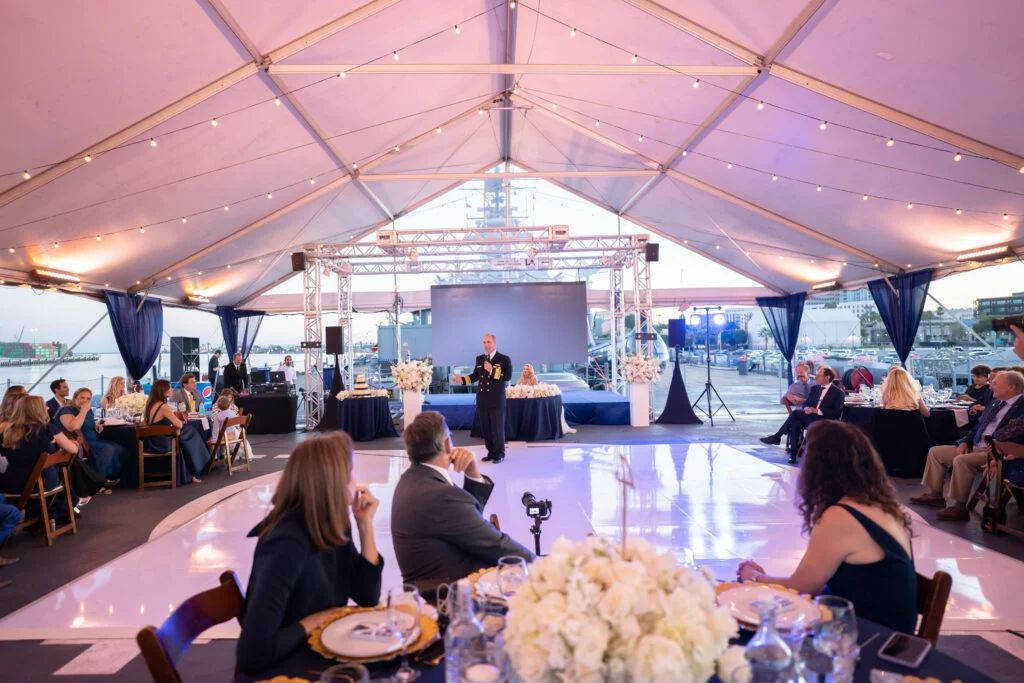 A man speaks onstage under a large white tent at a formal event venue, with guests seated at decorated tables and city buildings visible in the background.