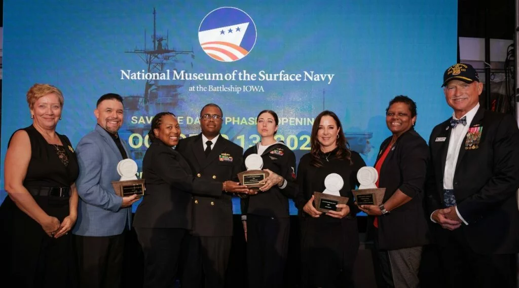 Eight people stand onstage, some in military uniforms, holding awards in front of a screen that reads National Museum of the Surface Navy at the Battleship IOWA. Smiling and posing for the camera, they embody a spirit of freedom and service honored by this distinguished recognition. Eight people stand onstage, some in military uniforms, holding awards in front of a screen that reads National Museum of the Surface Navy at the Battleship IOWA. Smiling and posing for the camera, they embody a spirit of freedom and service honored by this distinguished recognition.