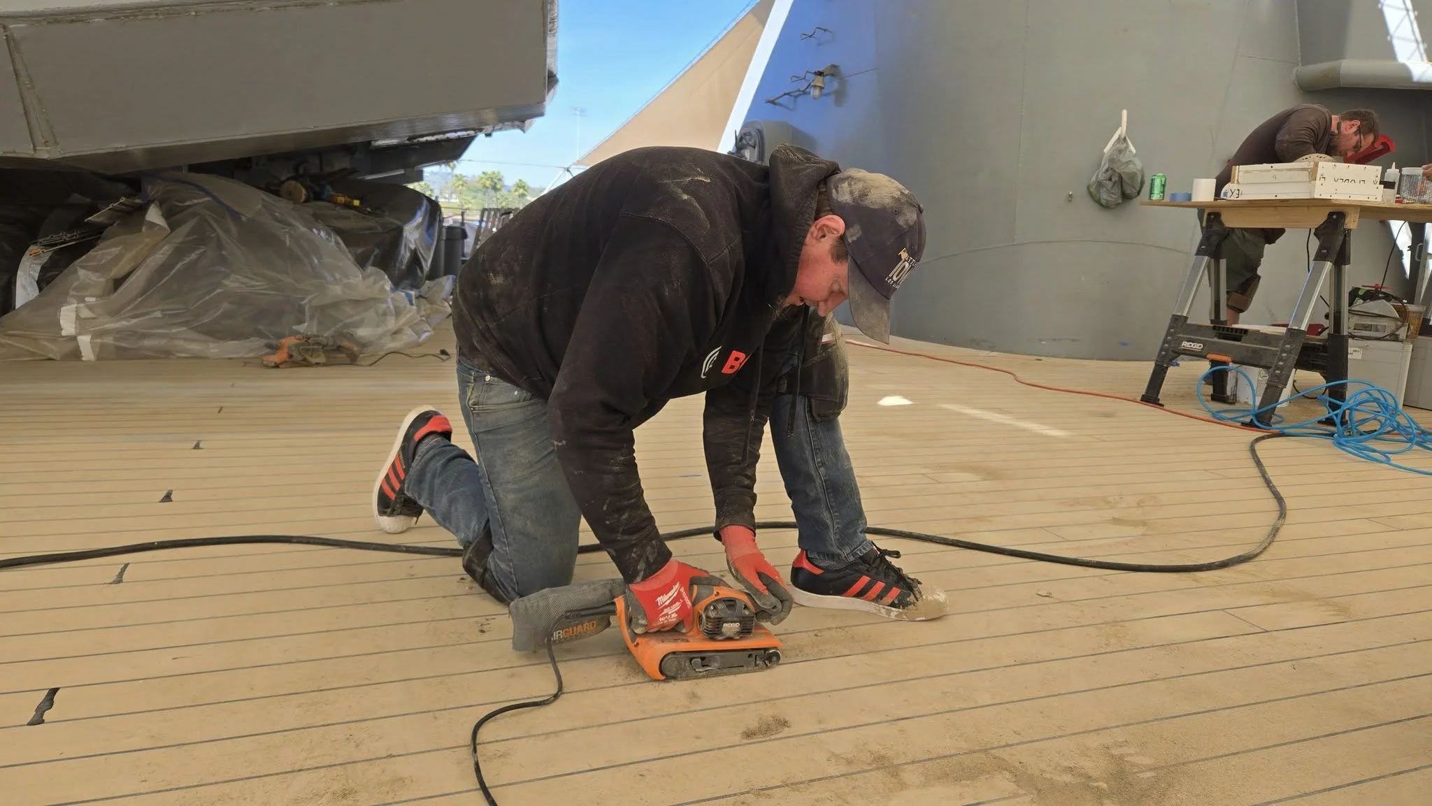 A person in a black hoodie and red gloves kneels on a wooden deck, sanding the surface with a power tool, much like volunteers restoring the historic battleship in Iowa. Tools, cords, and other equipment are scattered nearby.
