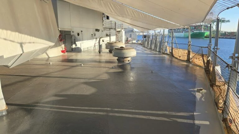 View of a battleship’s deck with metal vents, safety netting along the edge, and a canopy overhead; the industrial port of Los Angeles and a container ship are visible in the background near the water. View of a battleship’s deck with metal vents, safety netting along the edge, and a canopy overhead; the industrial port of Los Angeles and a container ship are visible in the background near the water.