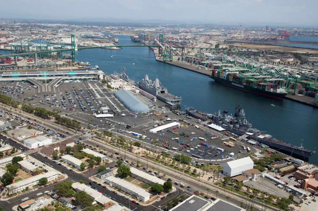 Aerial view of a busy port with cargo ships, shipping containers, cranes, parked cars, and naval ships docked along the waterfront, with industrial buildings and roads nearby, and a museum visible in the distance offering directions to visitors.