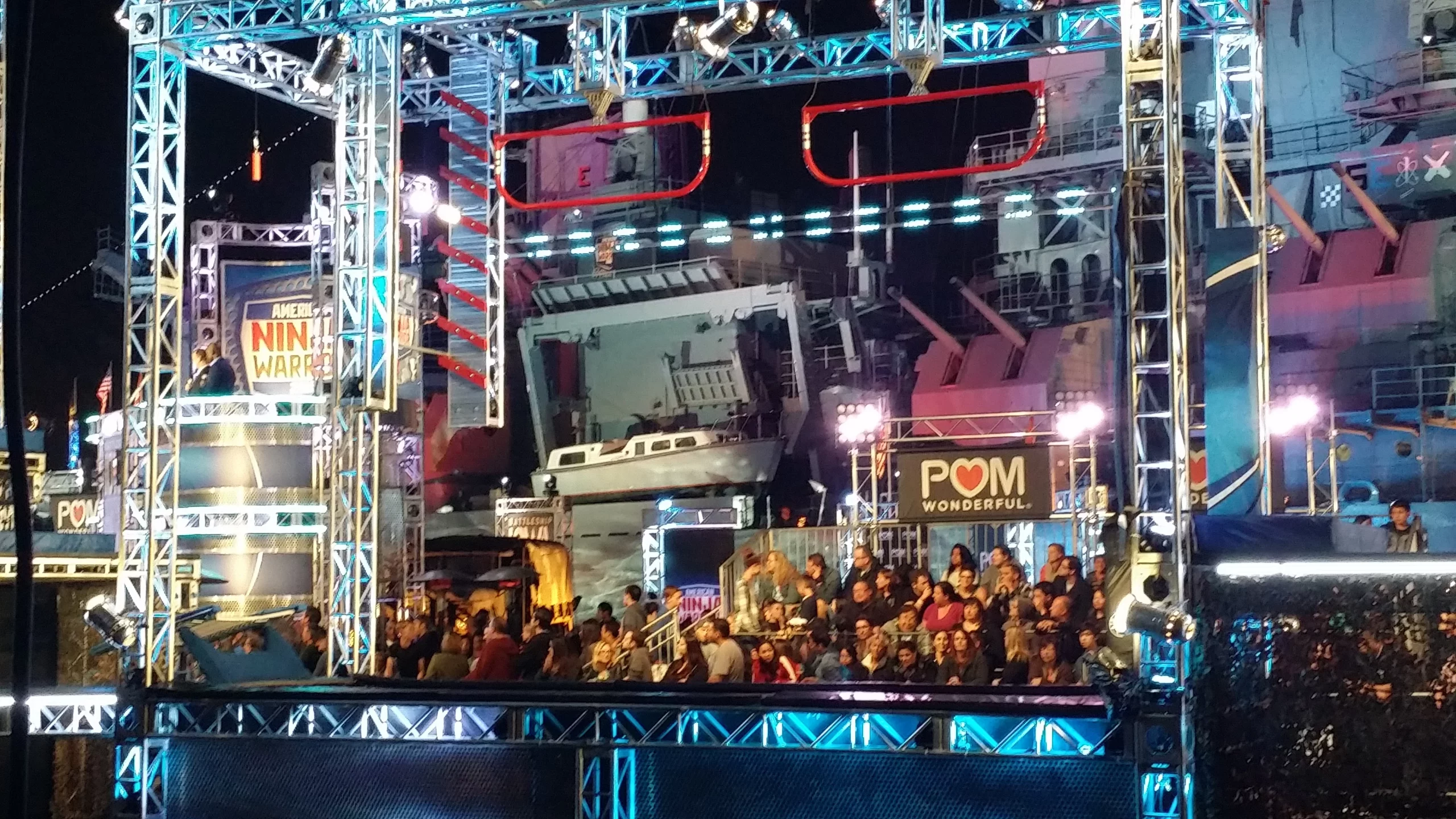 A brightly lit obstacle course set from American Ninja Warrior with a cheering audience in front of a backdrop featuring a large ship and Pom Wonderful ads.