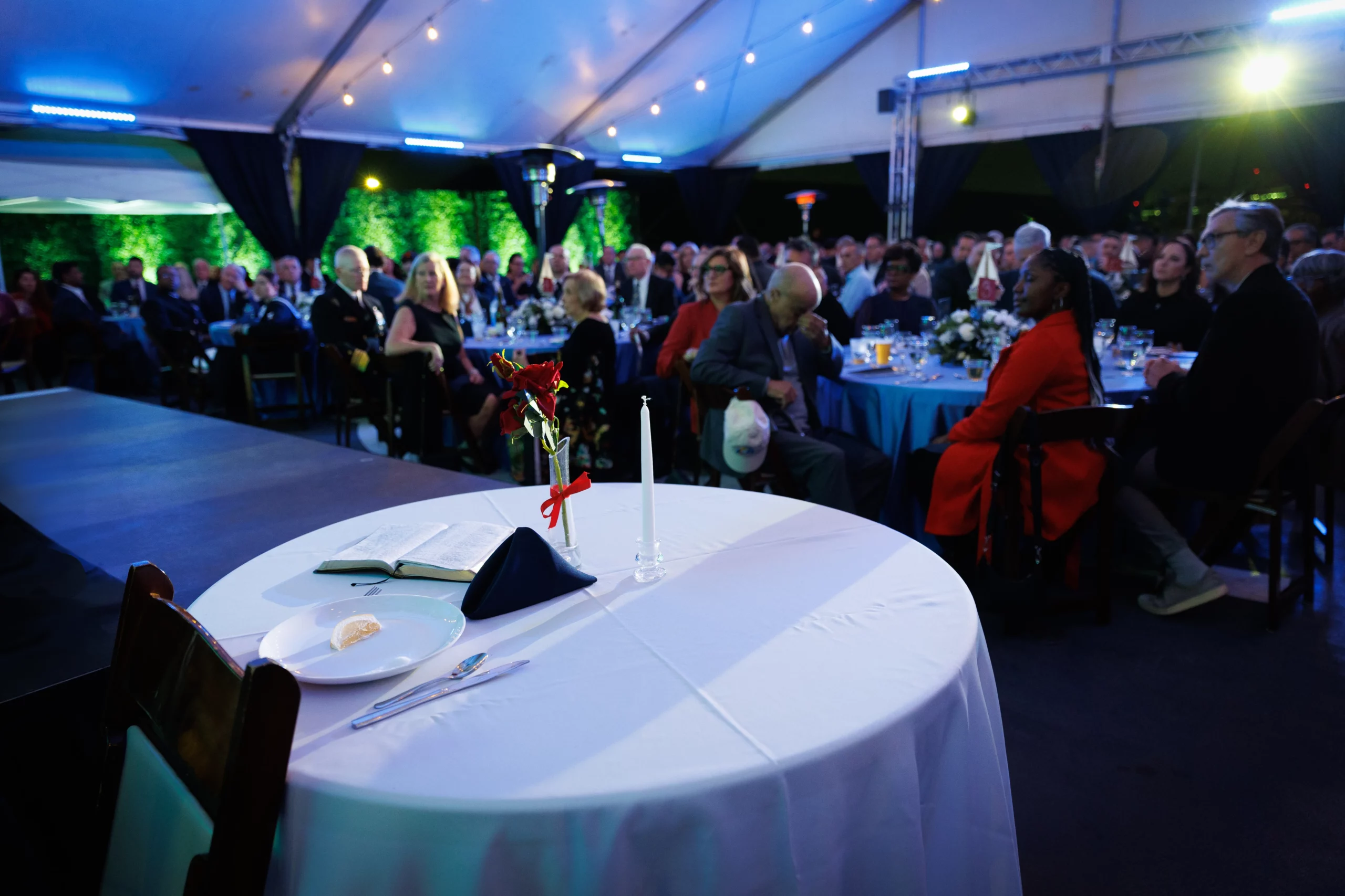 A round table with a white tablecloth, book, candle, and flower centerpiece stands in the foreground at a formal event for private events or groups, with many attendees seated and watching under a large tent.
