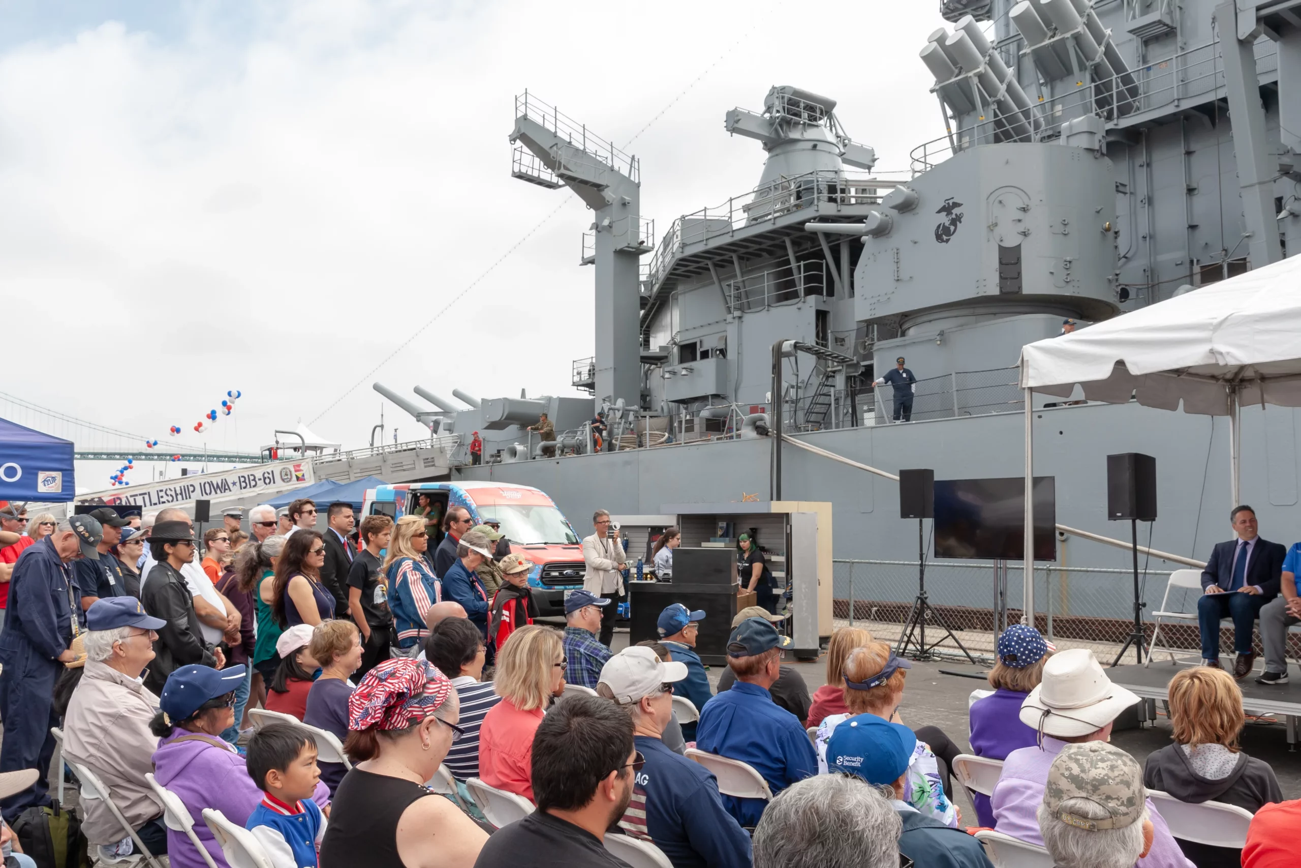 A crowd sits and stands listening to a speaker on stage near a large gray battleship. The event takes place outdoors, with people of various ages wearing casual clothing and hats.