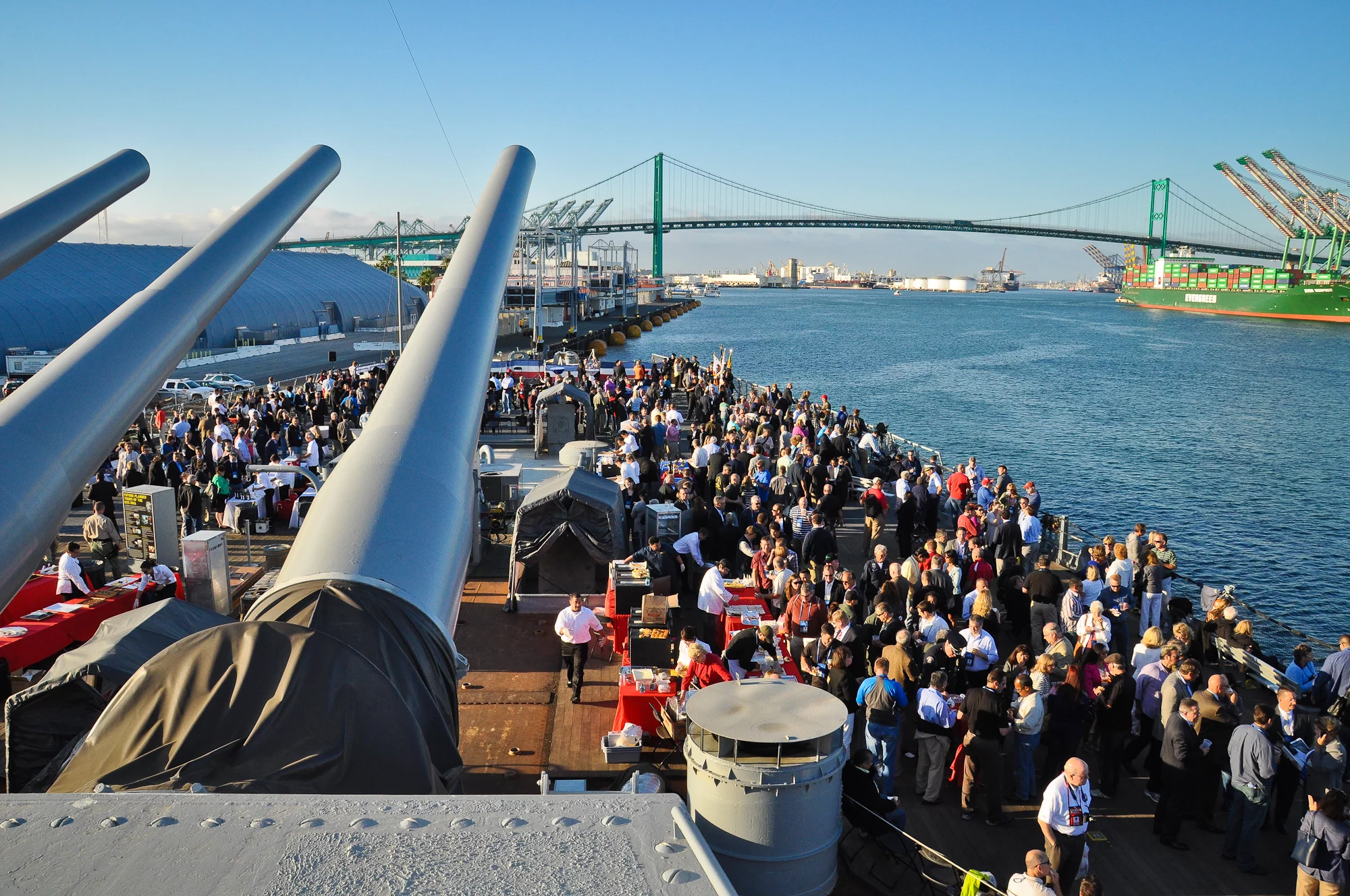 A large crowd gathers on the deck of a battleship docked by a port, with big cannons in the foreground and a suspension bridge, shipping containers, and cranes visible under a clear sky—an impressive setting ideal for private events or groups seeking a memorable experience.