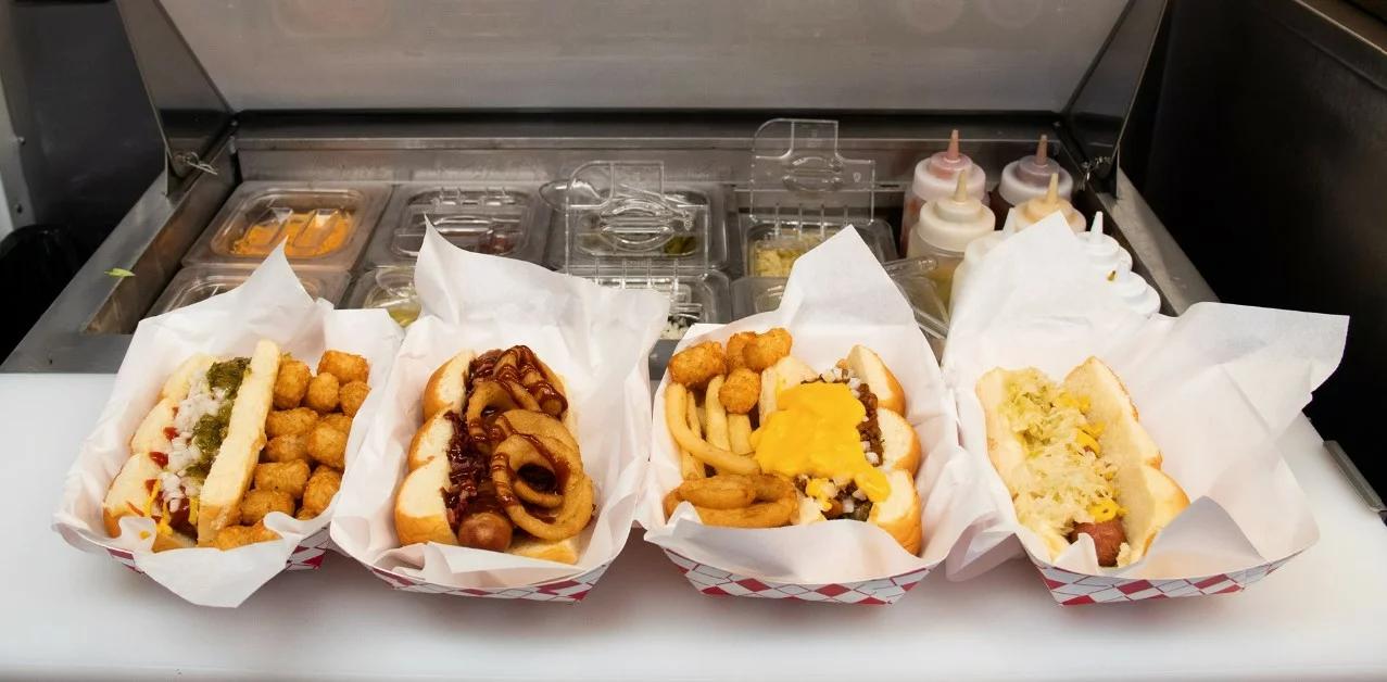 Four trays of loaded hot dogs with various toppings, served with tater tots, onion rings, and French fries, are displayed on a counter in front of a sandwich prep station with condiment bottles and ingredient trays, creating an experience reminiscent of visiting a food museum curated by Vicky.
