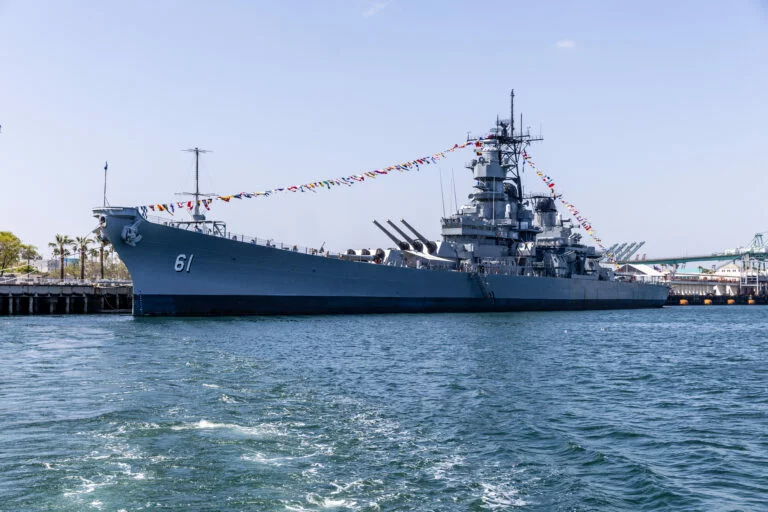 A large gray battleship numbered 61, now serving as a museum, is docked by a pier and decorated with colorful signal flags. The sky is clear and the water in the foreground is calm, inviting visitors to experience history aboard the impressive vessel.