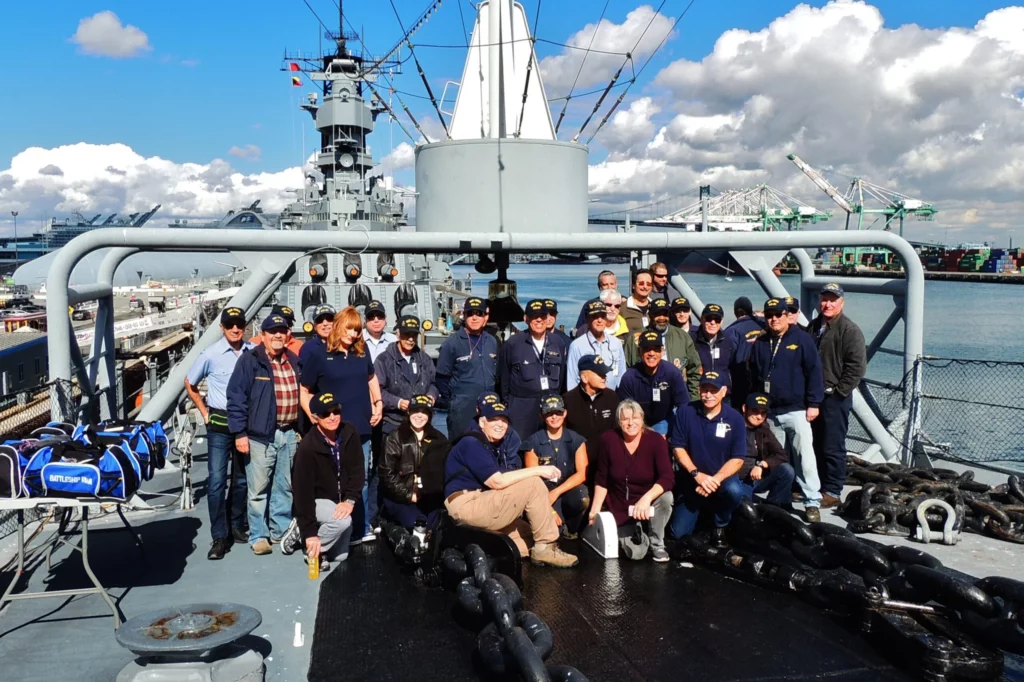 A group of people in navy hats and jackets pose on the deck of a museum ship under a blue sky with clouds and cranes visible in the background, inviting visitors in Los Angeles to explore and donate to support its preservation. A group of people in navy hats and jackets pose on the deck of a museum ship under a blue sky with clouds and cranes visible in the background, inviting visitors in Los Angeles to explore and donate to support its preservation.