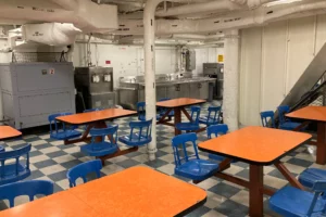 A cafeteria with orange tables, blue chairs, and a checkered floor, set in an industrial-looking room with exposed pipes and metal equipment in the background—an ideal event venue that offers a unique blend of vibrant style and urban atmosphere.