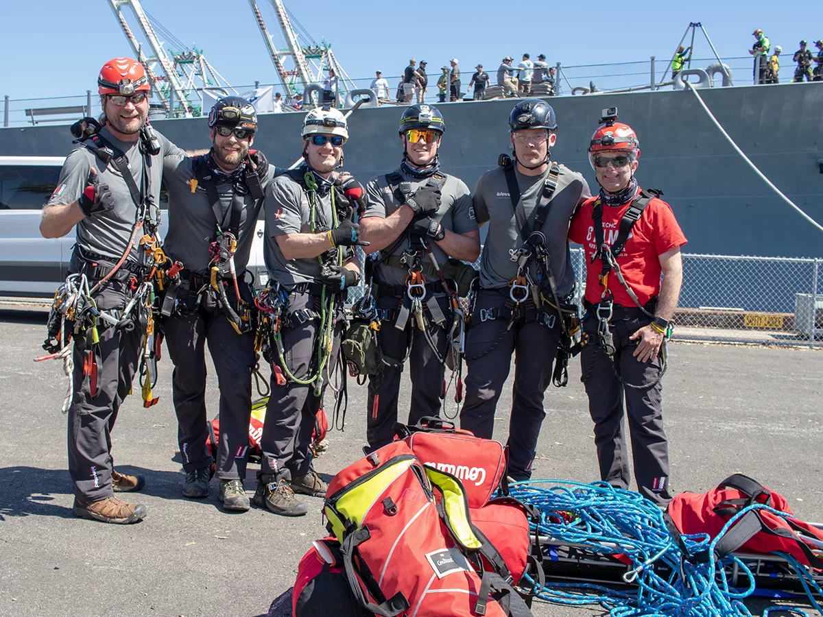 Six rescue workers, including several veterans, in helmets and harnesses stand together on a dock, posing for a photo. Bags and coiled ropes are on the ground in front of them. A large ship and several people are visible in the background.