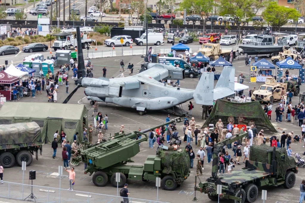 Aerial view of a crowded outdoor event at LA Fleet Week, displaying military vehicles, including a large aircraft, trucks, tents, and booths. People are walking around and observing the exhibits.
