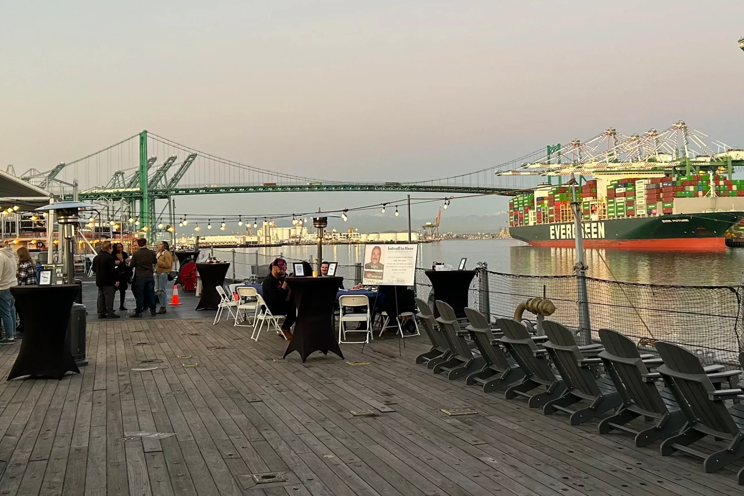 An outdoor event setup on a wooden pier overlooks a large cargo ship labeled EVERGREEN, with a suspension bridge and cranes in the background at sunset.