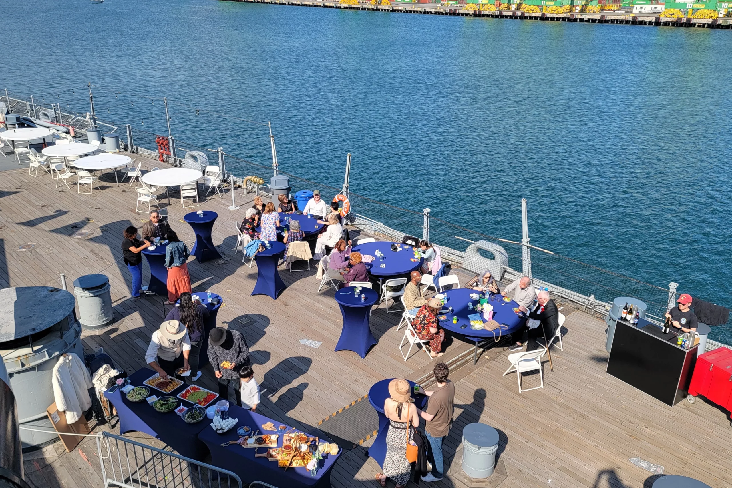 People gather on a docked ship for an outdoor event with blue tablecloths, food tables, and drinks, overlooking water and colorful shipping containers in the background.