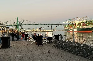 People gather on a waterfront wooden deck with tables and chairs. Cargo ships and cranes are visible in the background near a large green bridge at sunset.