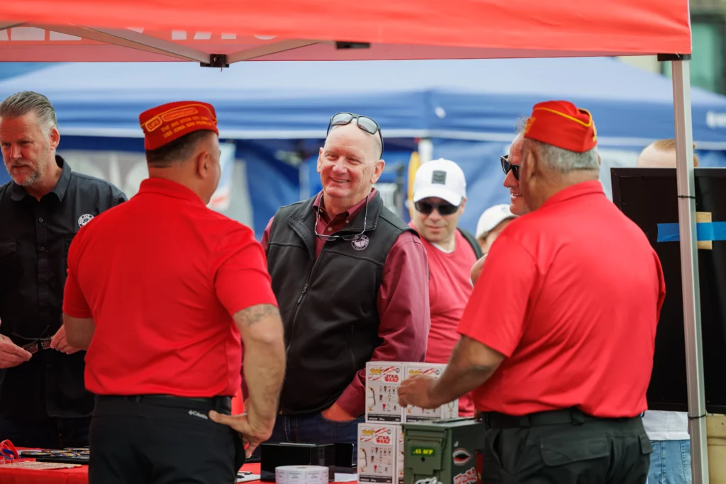 A group of men, some in red uniforms and hats, talk and laugh under a red canopy at an outdoor community event with tents in the background, highlighting the spirit of togetherness and shared programs.