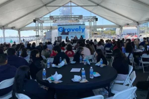 A group of kids sit at round tables under a large white tent, listening to speakers on a stage with a Gravely Experience banner. Bottles of water and snacks are on the tables, adding to the welcoming atmosphere of this Gravely event.