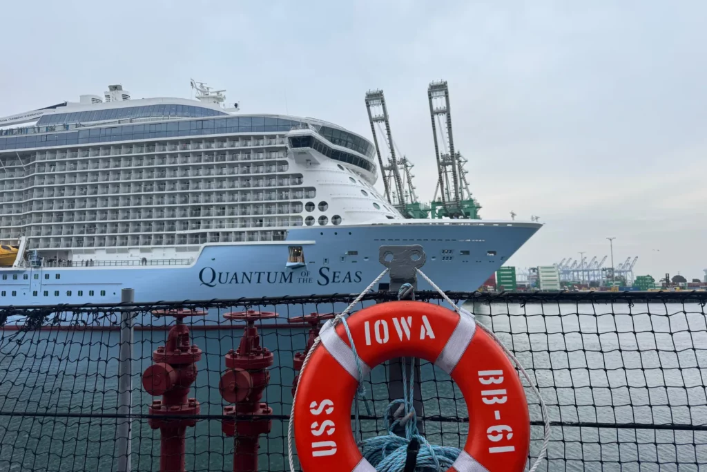 A large cruise ship named Quantum of the Seas is docked near the LA Cruise Terminal. In the foreground, a lifebuoy labeled USS IOWA BB-61 hangs on a metal fence above the water, capturing a scene characteristic of the bustling Los Angeles cruise port. A large cruise ship named Quantum of the Seas is docked near the LA Cruise Terminal. In the foreground, a lifebuoy labeled USS IOWA BB-61 hangs on a metal fence above the water, capturing a scene characteristic of the bustling Los Angeles cruise port.