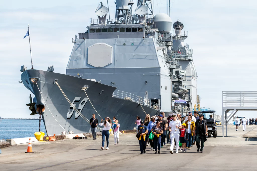 A large navy ship, the Battleship USS Iowa, is docked at a pier with a group of people, including families and children, walking away from the ship on a sunny day at the Los Angeles museum.