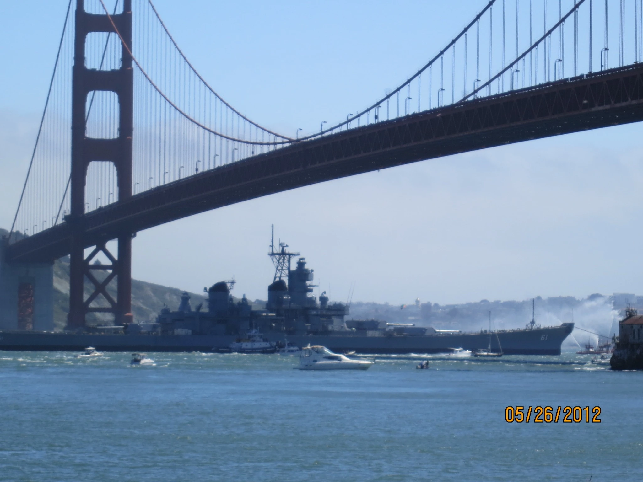 Battleship sails under the Golden Gate Bridge in San Francisco Bay, with several smaller boats nearby on a sunny day. The scene captures a classic display of naval presence reminiscent of The Broadside, set against the iconic backdrop. The date on the photo is May 26, 2012.