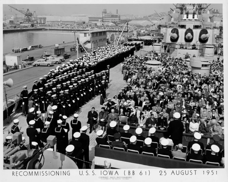 Black-and-white photo of a crowd seated on the deck of USS Iowa during a naval ceremony, with uniformed sailors standing in formation and ship structures visible. Text at bottom notes recommissioning on 25 August 1951.