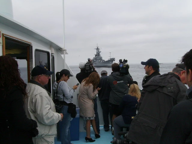 A group of people on a boat, many with cameras, photograph The Broadside—a large navy ship in the distance—under a cloudy sky.