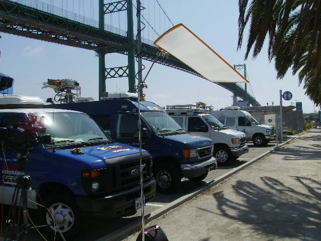 Several news vans with satellite equipment are parked by a sidewalk under a large suspension bridge on a sunny day, covering events for The Broadside. Cameras and lights are set up, with palm tree shadows visible in the foreground.