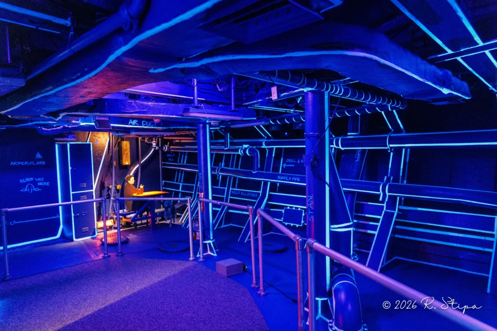 A futuristic, neon-lit room with blue and purple lights, metallic pipes, railings, and labeled panels, resembling a sci-fi spaceship interior aboard The Broadside. A workstation emits an orange glow in the background. A futuristic, neon-lit room with blue and purple lights, metallic pipes, railings, and labeled panels, resembling a sci-fi spaceship interior aboard The Broadside. A workstation emits an orange glow in the background.