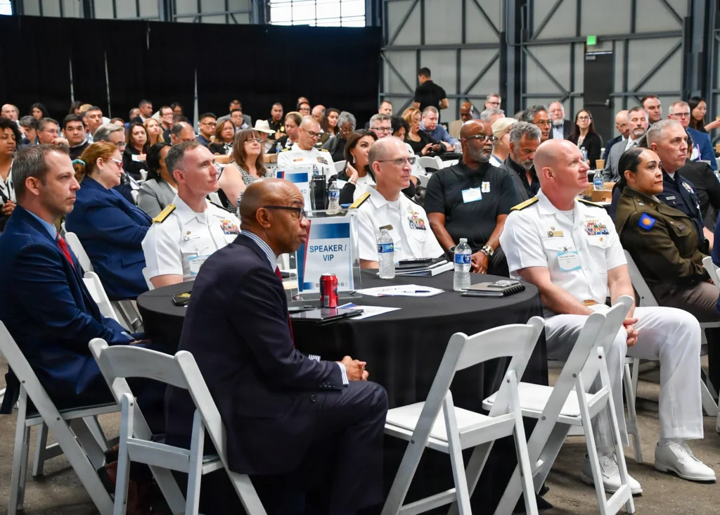 A diverse group of people, including military personnel in uniform and civilians, sit attentively at round tables during a conference or seminar in a large indoor venue.