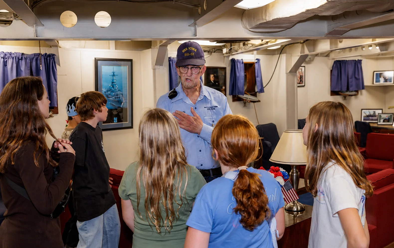 An older man in a navy hat speaks to a group of children inside a room with patriotic decorations, framed photos, and a small American flag visible on a table.
