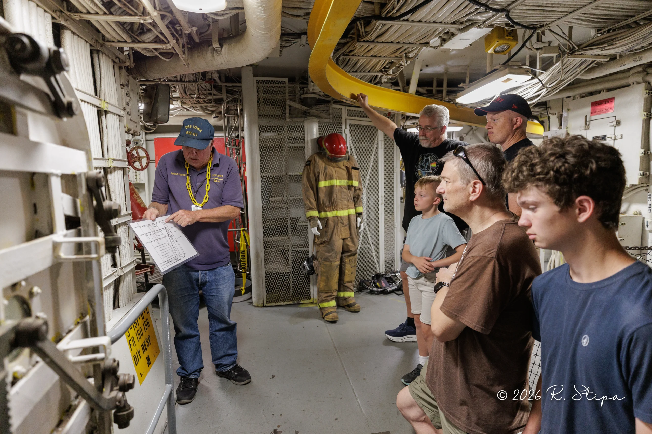 A man in a navy hat explains a checklist to a group of people, including children and a firefighter, inside a narrow, industrial corridor filled with pipes and wiring.