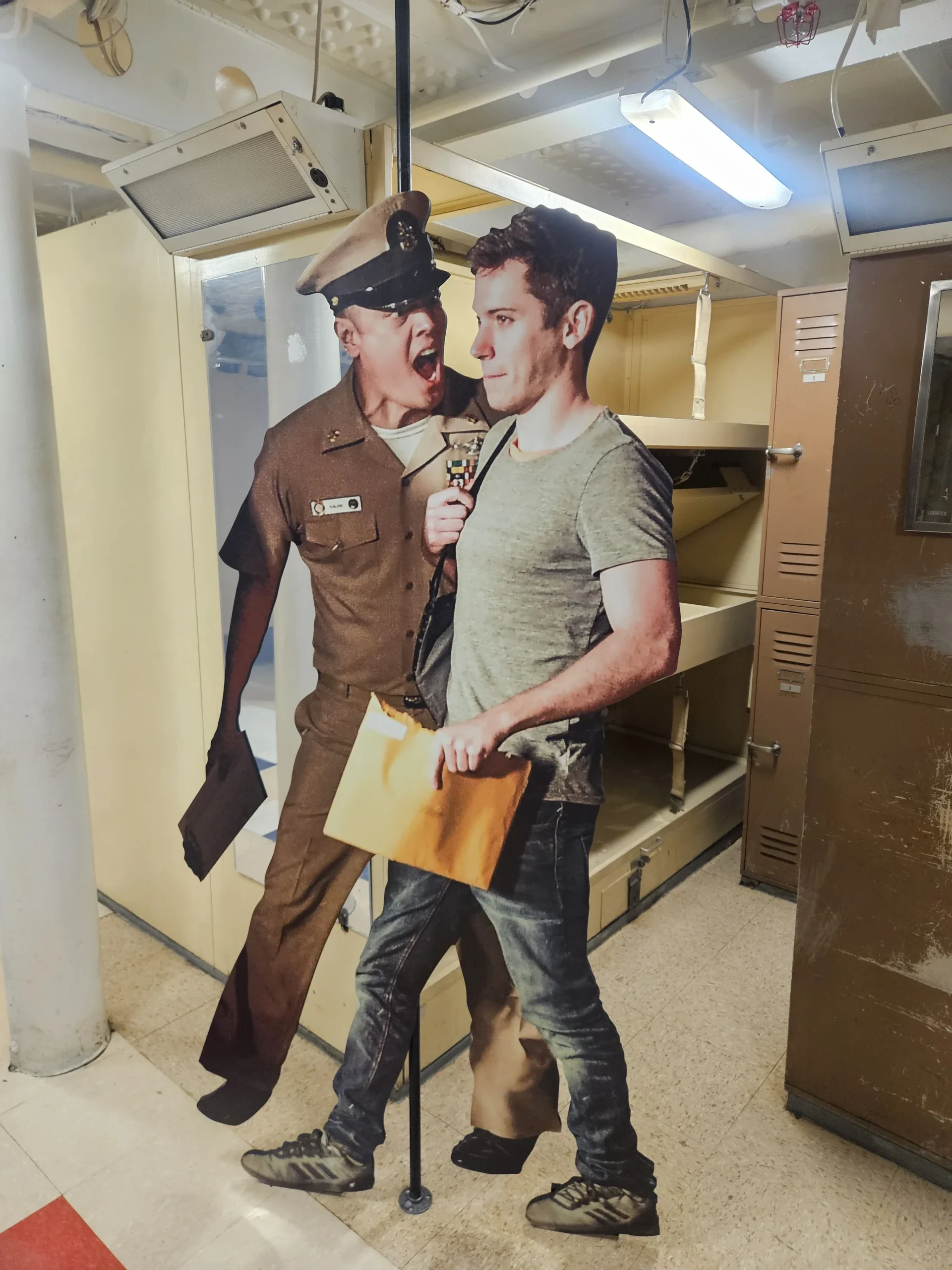 A cardboard cutout shows a uniformed officer yelling at a casually dressed man holding a large envelope, set in front of lockers and open shelving in an indoor space.