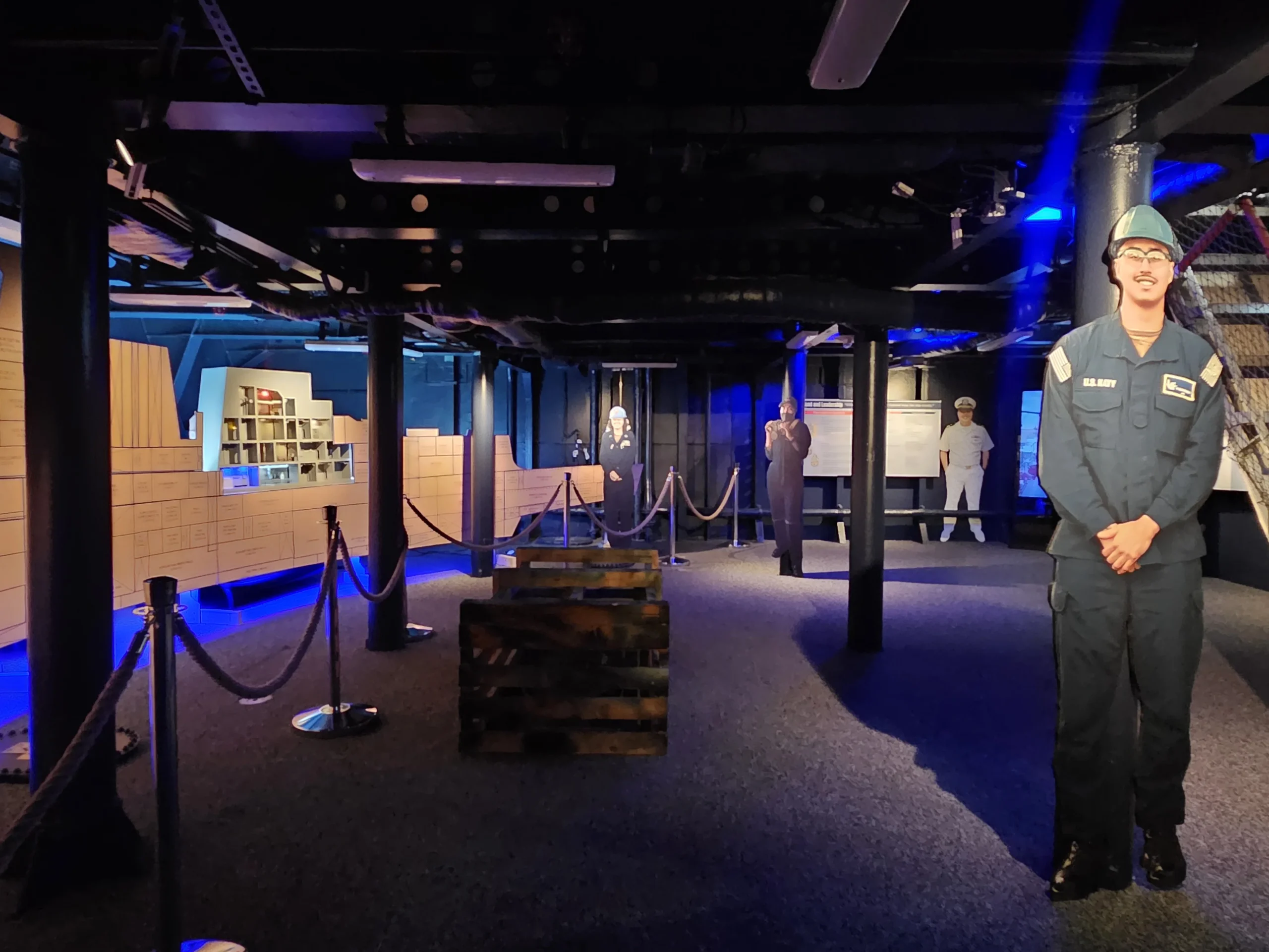 Exhibit with dim blue lighting, crates, rope barriers, life-size cutouts of people in uniforms, and a background resembling a ships interior and a library area.