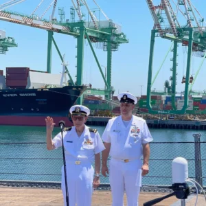 Chief Warrant Officer Al Canfield, USN (Ret.) and CPO David Canfield, USN (Ret.) Two naval officers in white uniforms stand by a microphone at a port, with large cranes and a cargo ship labeled Ever Shine in the background on a sunny day.