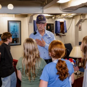 Volunteer Dale Chitwood with students An older man in a navy hat speaks to a group of children inside a room with naval decor, including ship photos and small American flags, as they listen attentively.