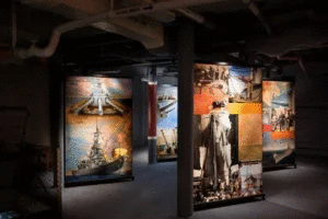 Dimly lit museum exhibit with large panels displaying historic photos and illustrations of battleships, Navy personnel, and World War II scenes, surrounded by pipes and industrial decor.