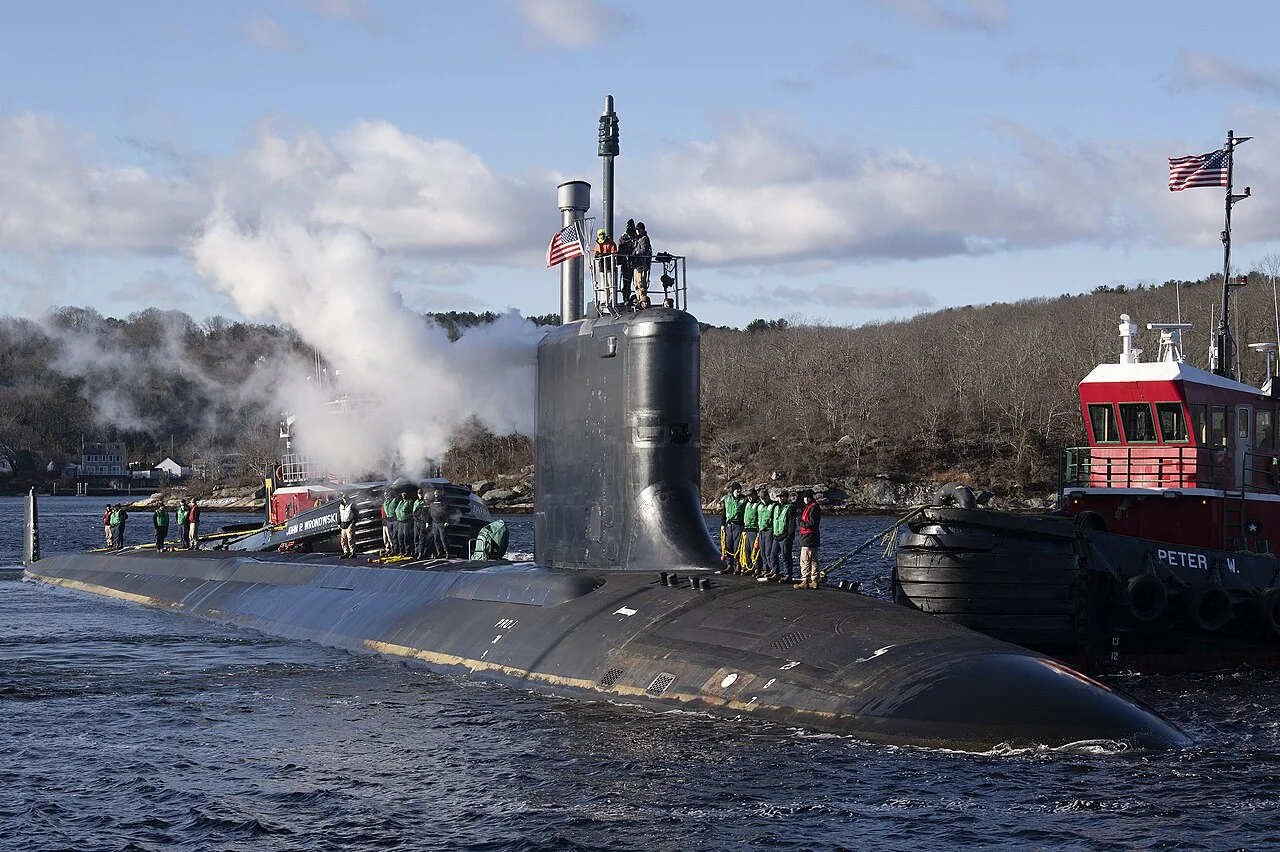 A submarine surfaces near the shore as crew members in green vests stand on deck. Steam rises from the sub, while tugboats assist. Trees and buildings line the background under a partly cloudy sky. USS Iowa SSN-797 delivery.