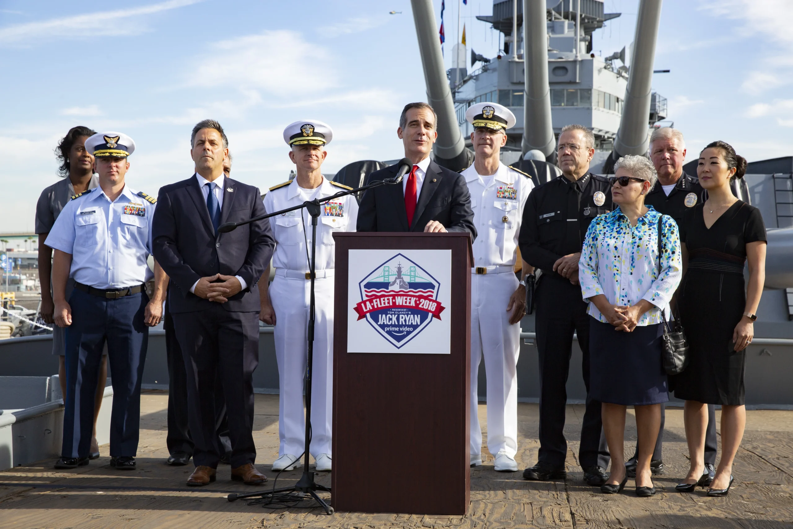 A group of people, including military officers and officials, stand on a ship deck behind a podium labeled LA Fleet Week 2022 Jack Pan, with a cityscape and battleship visible in the background.