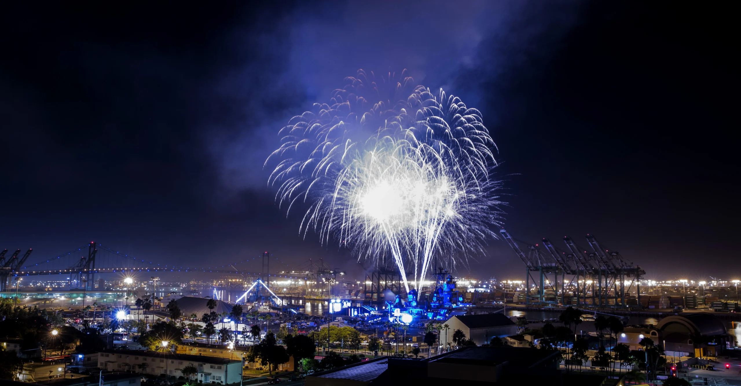 Fireworks burst in the night sky over a brightly lit harbor with cranes, ships, and buildings visible below, creating a festive and lively atmosphere.