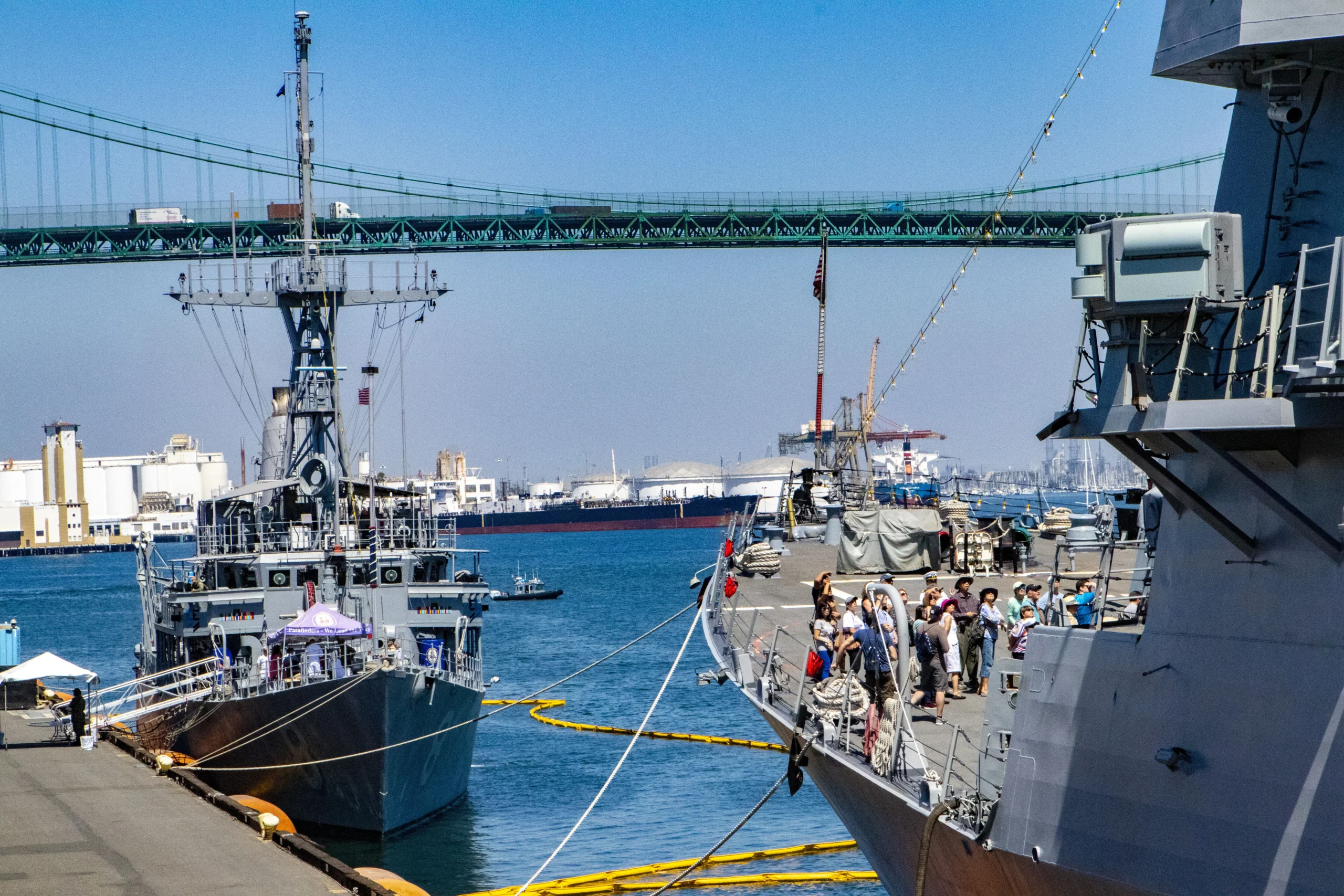 Two navy ships docked at a harbor with people standing on one deck. A bridge and industrial buildings are visible in the background under a clear blue sky.