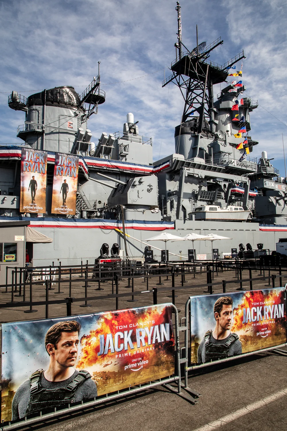 A battleship decorated with banners and posters promoting the TV show Jack Ryan featuring the main character, with barricades displaying the same promotional images in the foreground.