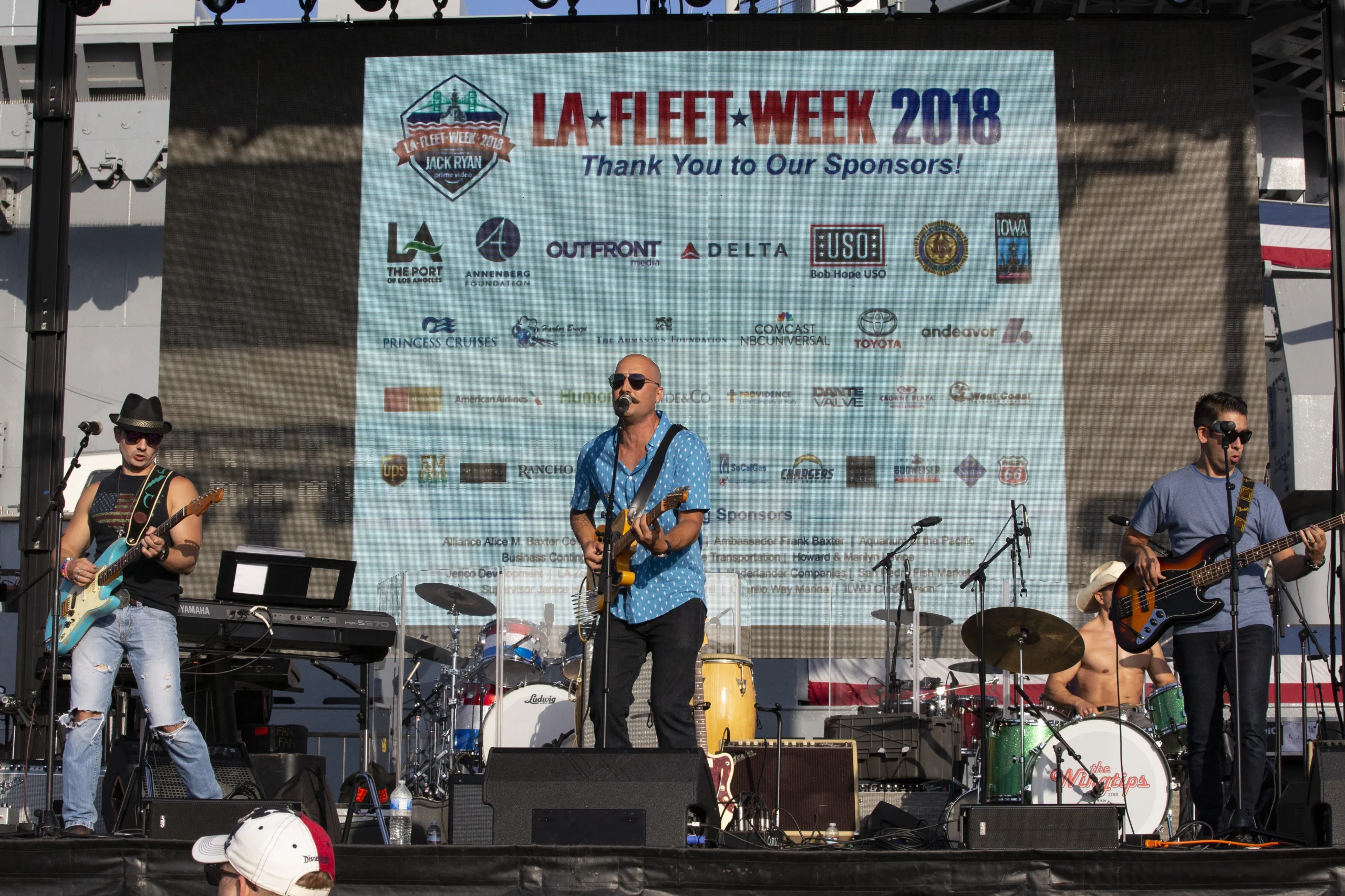 A band performs onstage at LA Fleet Week 2018, with musicians playing guitar, keyboard, and drums in front of a large event sponsor banner.