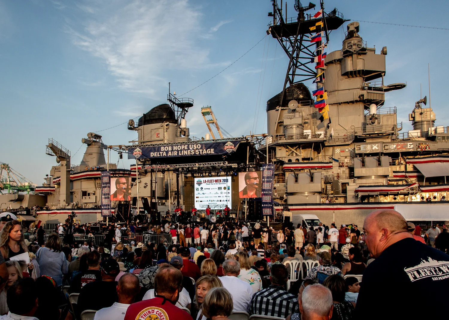 A large crowd attends an outdoor event on a battleship, facing a stage with screens and banners. Colorful flags decorate the ship, and people are seated and standing, enjoying the show under a clear sky.