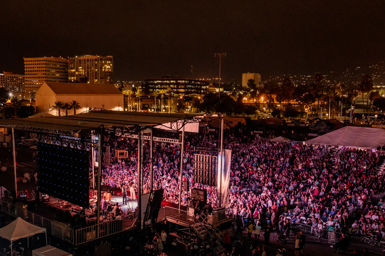 A large outdoor concert at night with a stage lit up and a crowd of people gathered. City buildings and lights are visible in the background.