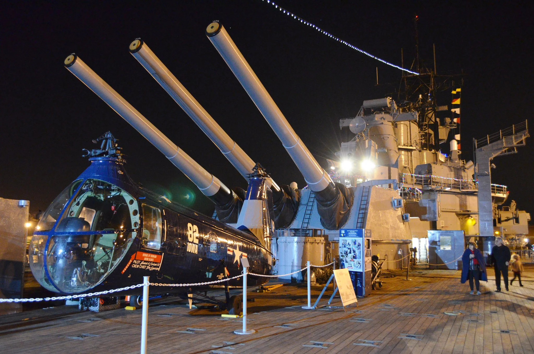 A helicopter and three large naval gun turrets are displayed on the deck of a battleship at night, with lights illuminating the area and people walking nearby.