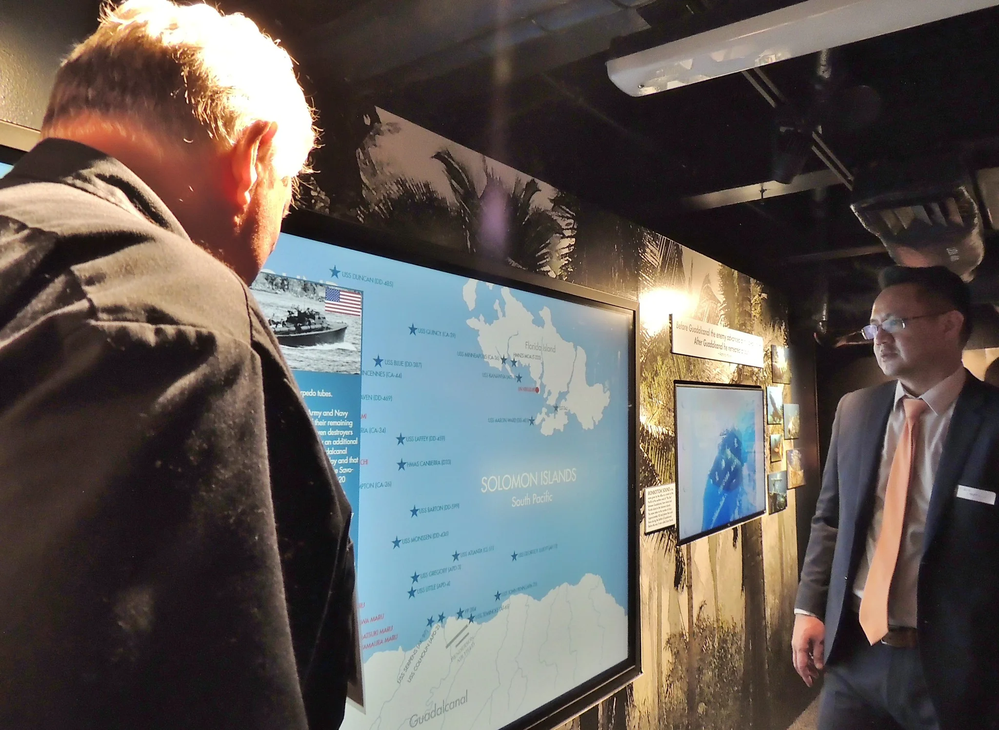 Two men in suits look at an illuminated exhibit display about the Solomon Islands, featuring maps and photos, in a dimly lit museum or gallery space.