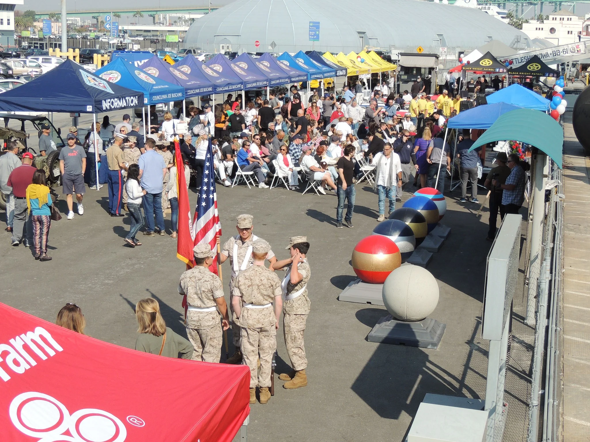 A crowded outdoor event features military personnel holding an American flag, booths with colorful awnings, seated and standing attendees, and large decorative spheres along a fence.