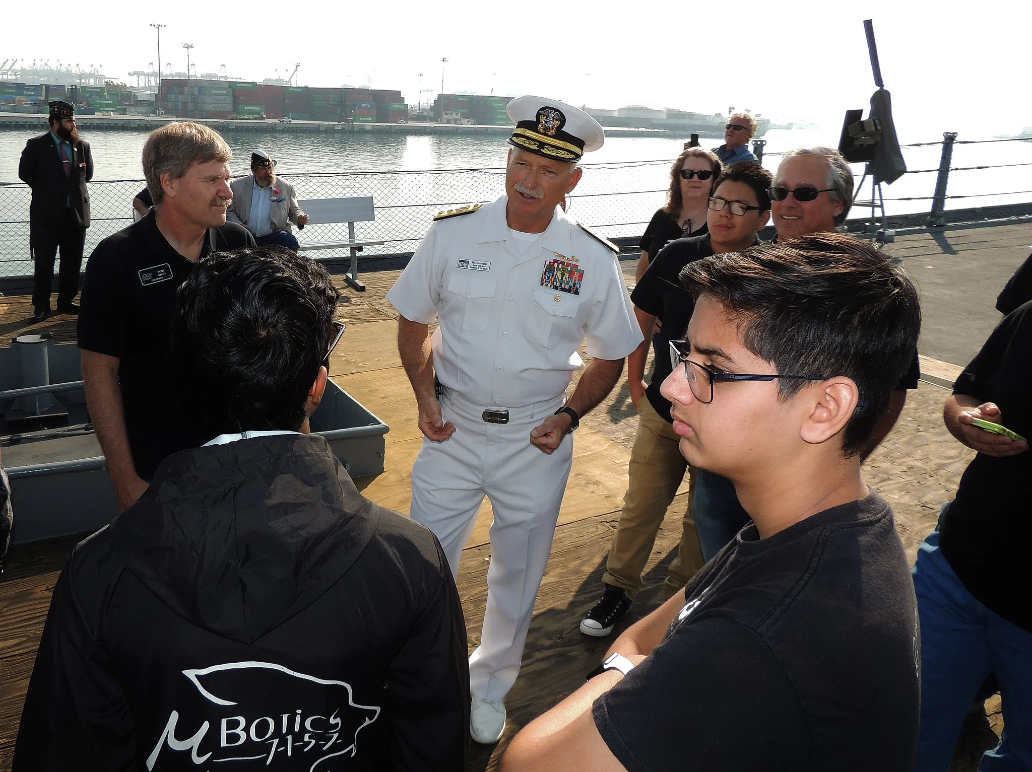 A naval officer in a white uniform talks with a group of people, including teens and adults, on a ships deck with industrial cranes and containers visible in the background.