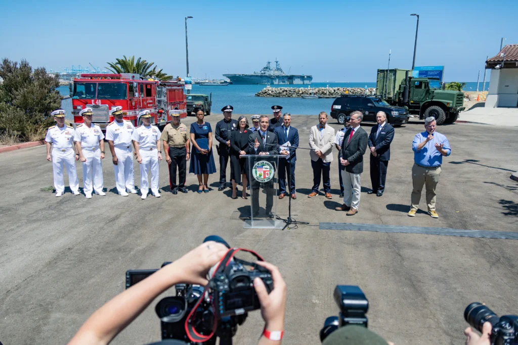 A group of officials and military personnel stand at a podium near the waterfront, with fire trucks, military vehicles, and a naval ship in the background. Photographers capture the scene from the foreground.